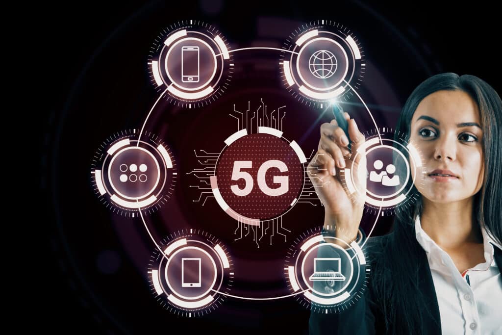 Uma mulher de negócios, com cabelos escuros e vestindo um blazer preto, interage com uma interface digital futurista em um fundo escuro. Ela utiliza uma caneta digital para tocar em um dos ícones de um diagrama circular que tem o destaque "5G" ao centro.