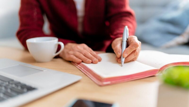 uma mulher escrevendo em um caderno em uma mesa, e ao seu lado uma xicara de café