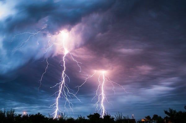 Raios intensos cruzam o céu durante uma tempestade, iluminando nuvens escuras acima de uma área com árvores e construções.
