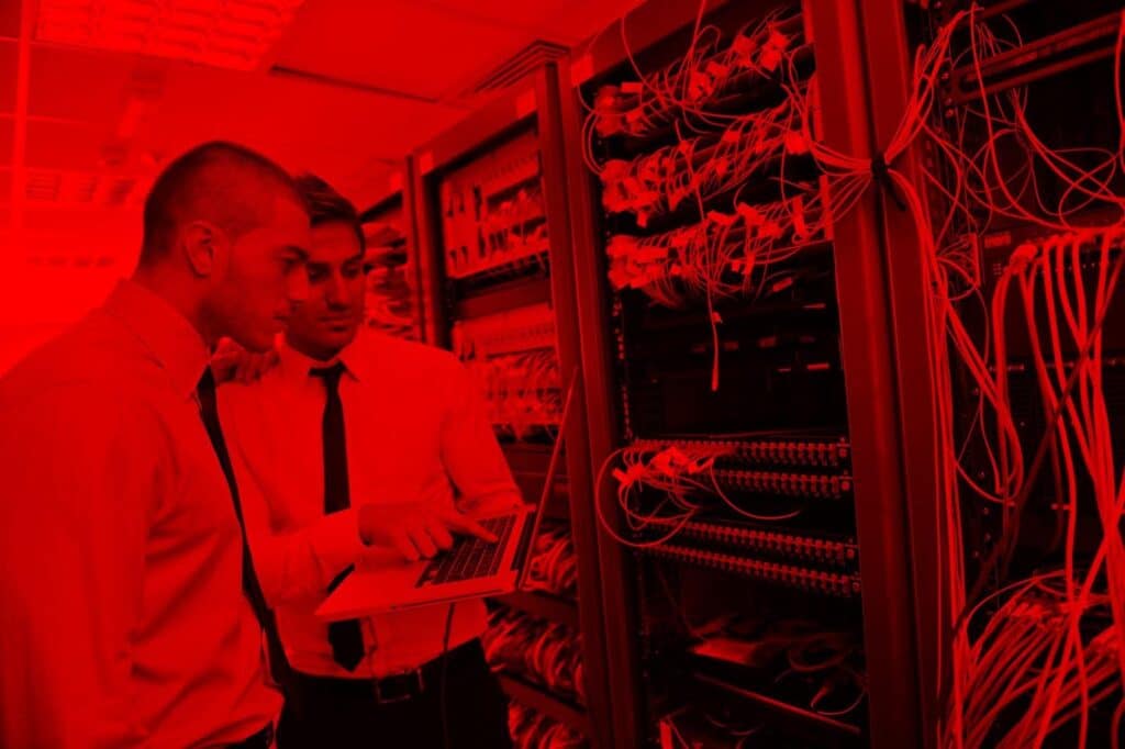 Dois homens de terno e gravata estão em uma sala de servidores, cercados por cabos e equipamentos de rede. Um deles segura um notebook aberto, enquanto ambos observam atentamente a tela, aparentemente discutindo ou analisando informações técnicas. Toda a cena está coberta por um filtro vermelho intenso, reforçando a atmosfera tecnológica.