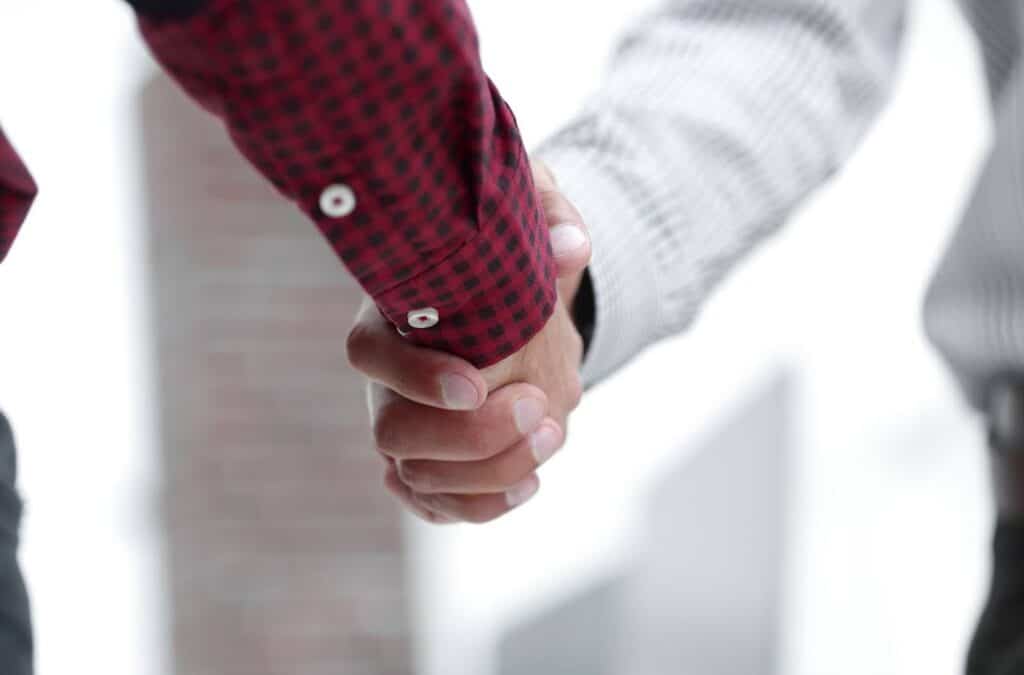Esta imagem é um close-up de duas pessoas apertando as mãos. A pessoa da esquerda está usando uma camisa de flanela vermelha com um padrão xadrez preto e botões brancos. A pessoa da direita está usando uma camisa cinza com listras finas, que parecem ser de um terno ou roupa social. O foco principal da imagem está nas mãos, que estão firmemente unidas. O fundo está desfocado e claro, sugerindo um ambiente ao ar livre ou com muita luz natural, talvez um prédio de escritórios. A imagem evoca a ideia de um acordo, parceria ou cumprimento profissional.