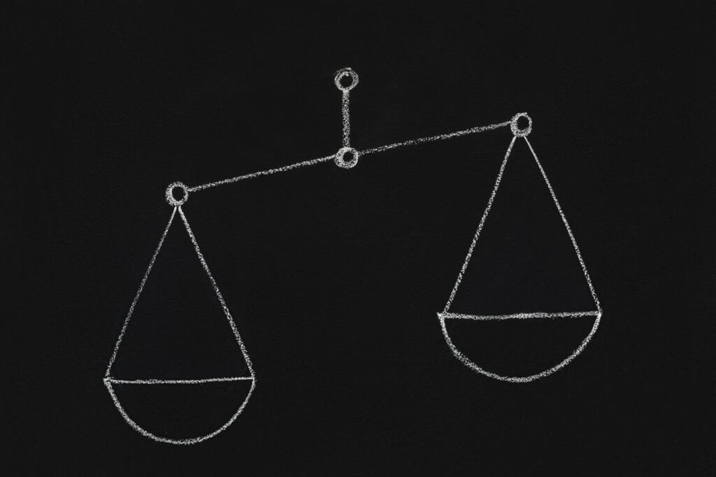 A imagem mostra um desenho de uma balança feita com giz, com o fundo preto. As duas tigelas da balança estão desenhadas de forma simétrica, penduradas em um eixo central. O desenho é simples, mas transmite a ideia de equilíbrio e justiça, com uma linha fina conectando as duas partes da balança.