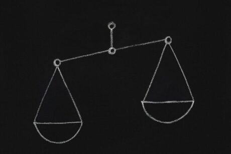 A imagem mostra um desenho de uma balança feita com giz, com o fundo preto. As duas tigelas da balança estão desenhadas de forma simétrica, penduradas em um eixo central. O desenho é simples, mas transmite a ideia de equilíbrio e justiça, com uma linha fina conectando as duas partes da balança.