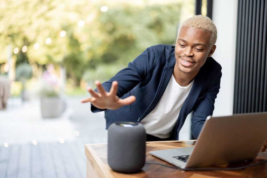 Uma fotografia horizontal de um homem negro, com cabelo descolorido e loiro, vestindo uma camisa branca e um blazer azul-marinho. Ele está em uma área externa, possivelmente uma varanda ou pátio, com a mão direita estendida para um alto-falante cilíndrico e cinza que está em uma mesa de madeira. O homem está sorrindo e olhando para o alto-falante, parecendo interagir com ele. Há um laptop aberto e prateado à sua direita e um fundo com folhagem verde embaçada e luzes de jardim.