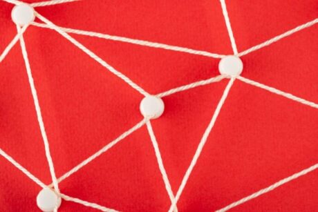 Uma imagem de um fundo vermelho vibrante, com vários pontos circulares brancos unidos por linhas brancas de barbante ou corda. Os pontos brancos são colocados aleatoriamente, e as linhas brancas se cruzam, formando uma variedade de triângulos de tamanhos diferentes, representando uma rede de conexões.
