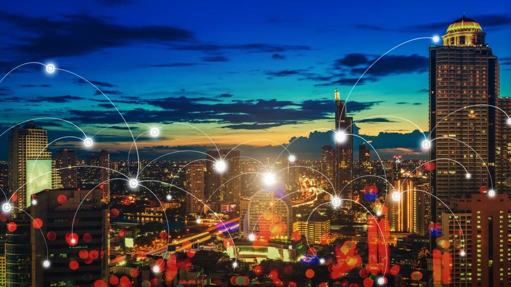 A imagem mostra uma vista panorâmica de uma cidade à noite, com vários prédios iluminados. O céu ao fundo tem tons de azul e laranja, sugerindo o fim do dia. Sobre a cidade, há linhas conectando pontos brilhantes, representando uma rede de conexões, como uma representação visual de dados ou comunicação digital. As linhas e os pontos criam um efeito de tecnologia, com foco nas conexões entre as áreas urbanas. As luzes nos prédios e nas ruas refletem a vida noturna da cidade, criando um cenário vibrante.