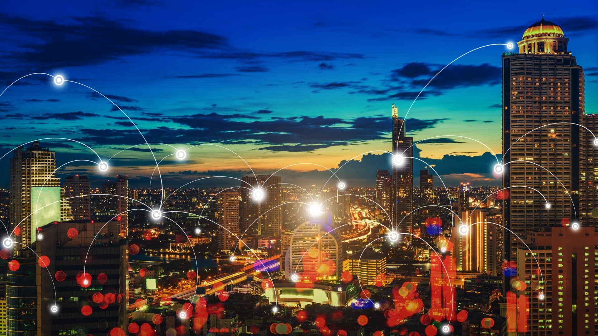 A imagem mostra uma vista panorâmica de uma cidade à noite, com vários prédios iluminados. O céu ao fundo tem tons de azul e laranja, sugerindo o fim do dia. Sobre a cidade, há linhas conectando pontos brilhantes, representando uma rede de conexões, como uma representação visual de dados ou comunicação digital. As linhas e os pontos criam um efeito de tecnologia, com foco nas conexões entre as áreas urbanas. As luzes nos prédios e nas ruas refletem a vida noturna da cidade, criando um cenário vibrante.