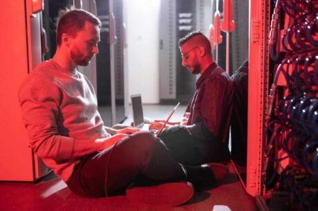A imagem mostra dois profissionais trabalhando em um ambiente de data center. Eles estão sentados no chão, com laptops, em meio a servidores e cabos. A iluminação vermelha cria uma atmosfera intensa e tecnológica, enquanto os dois indivíduos parecem focados em suas tarefas, ajustando ou monitorando os sistemas.