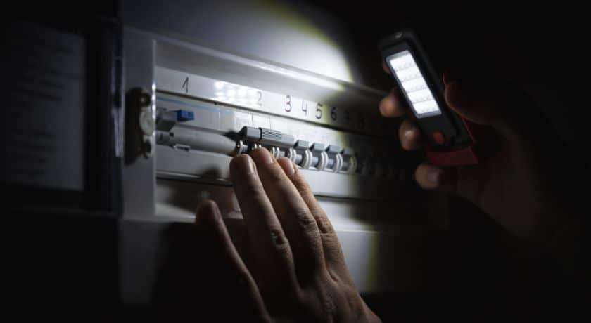 Um ambiente escuro, provavelmente devido a uma falha de energia (blecaute). Uma mão humana está na frente de um quadro de distribuição elétrico (painel de disjuntores), tentando manusear um dos disjuntores. A única fonte de luz visível é o feixe brilhante de uma lanterna LED