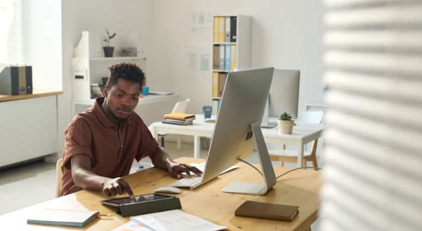 um homem negro jovem trabalhando em um escritório moderno, bem iluminado e organizado. Ele está sentado à mesa de madeira, concentrado, enquanto utiliza um tablet com a mão esquerda e tem um computador desktop à sua frente