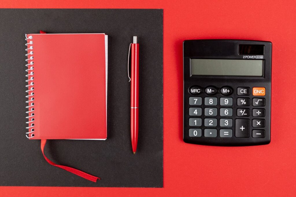 Sobre um fundo dividido entre vermelho e preto, há um caderno espiral vermelho fechado, posicionado à esquerda, com uma fita marcadora visível. Ao lado do caderno, aparece uma caneta vermelha alinhada verticalmente. À direita, há uma calculadora preta com visor digital e botões numéricos, organizada de forma simétrica, sugerindo um ambiente de planejamento ou controle financeiro.